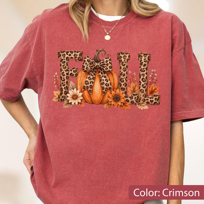 May include: A crimson short-sleeve t-shirt featuring a graphic print of the word FALL. The letters are filled with a leopard print pattern. A central orange pumpkin with a leopard print bow sits between the letters, surrounded by autumn flowers.