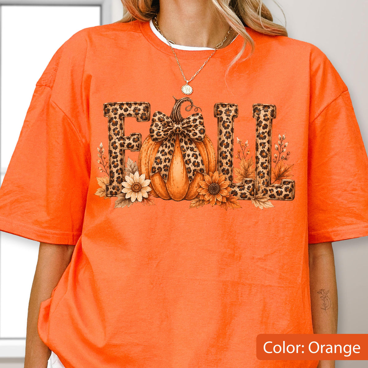 May include: An orange t-shirt featuring a graphic print of the word FALL in a leopard print pattern. The letter A is replaced by an orange pumpkin with a leopard print bow. The design includes sunflowers and wheat stalks at the base.