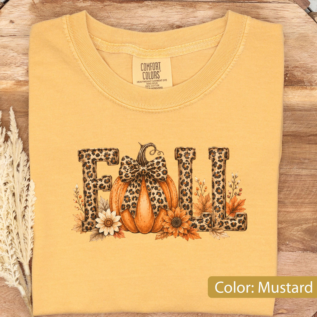 May include: A mustard yellow t-shirt featuring the word FALL in a leopard print pattern. The letter A is replaced by an orange pumpkin with a leopard print bow. The design includes small sunflowers and autumn leaves around the base of the letters.