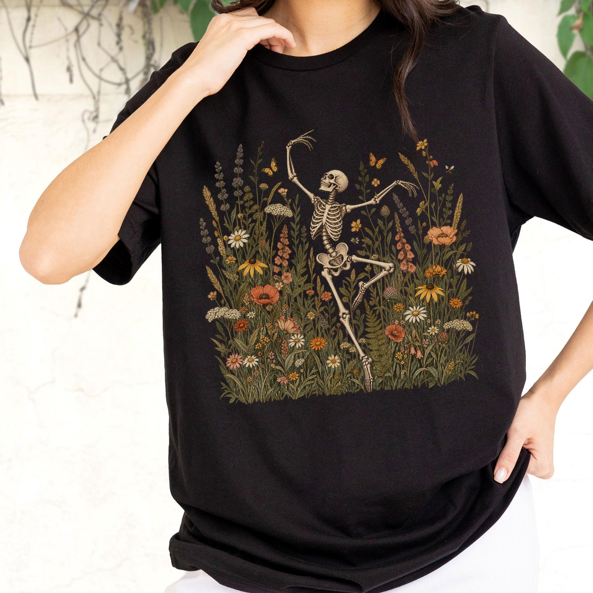 May include: A black short-sleeved t-shirt featuring a graphic print of a human skeleton dancing amidst a field of colorful wildflowers, including orange poppies, white daisies, and green foliage, with small butterflies fluttering above.