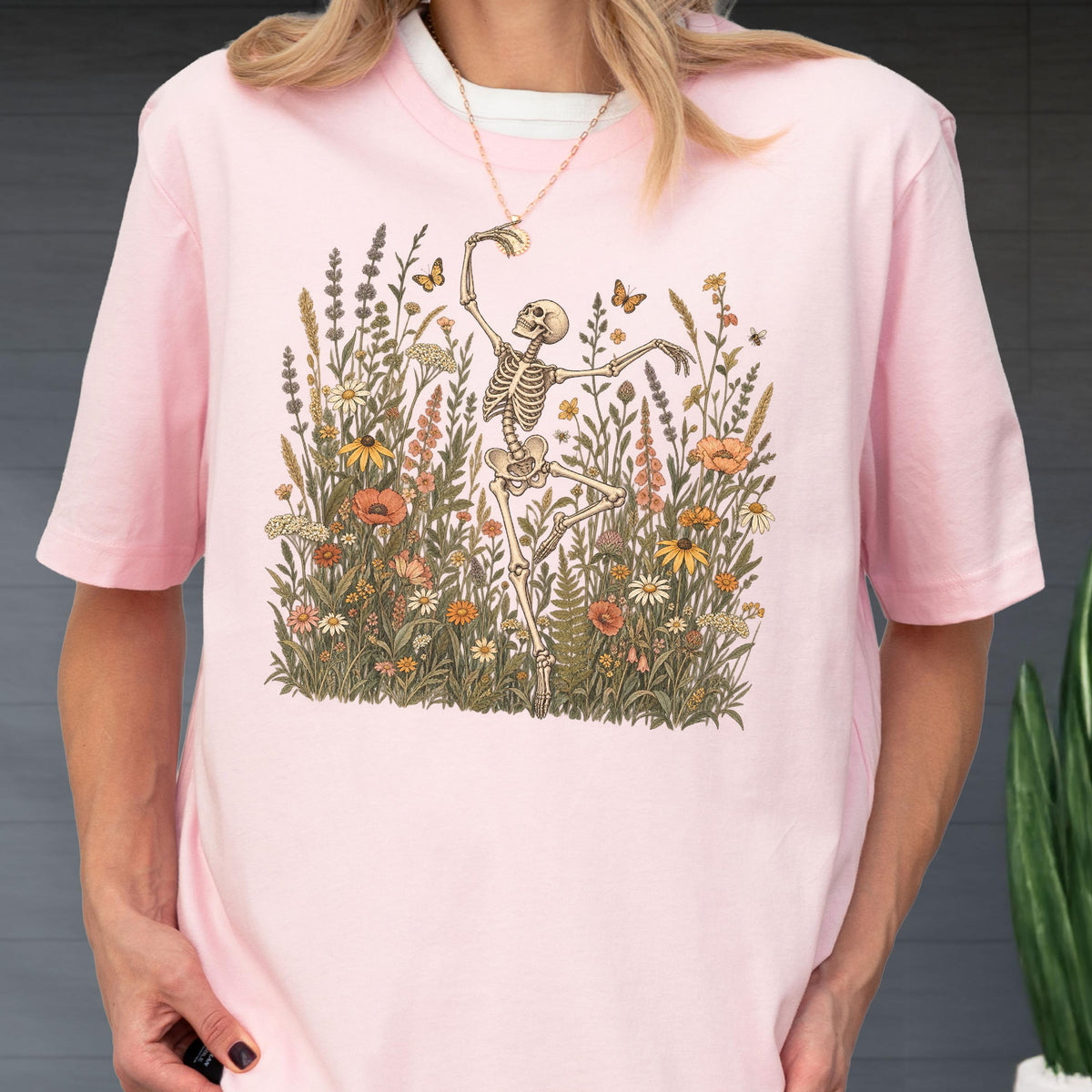 May include: A light pink short-sleeve t-shirt featuring a graphic print of a skeleton dancing amidst a field of colorful wildflowers, tall grasses, and small butterflies. The design is centered on the chest, showcasing detailed botanical illustrations.