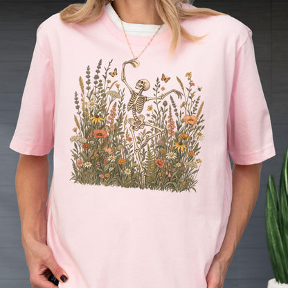 May include: A light pink short-sleeve t-shirt featuring a graphic print of a skeleton dancing amidst a field of colorful wildflowers, tall grasses, and small butterflies. The design is centered on the chest, showcasing detailed botanical illustrations.