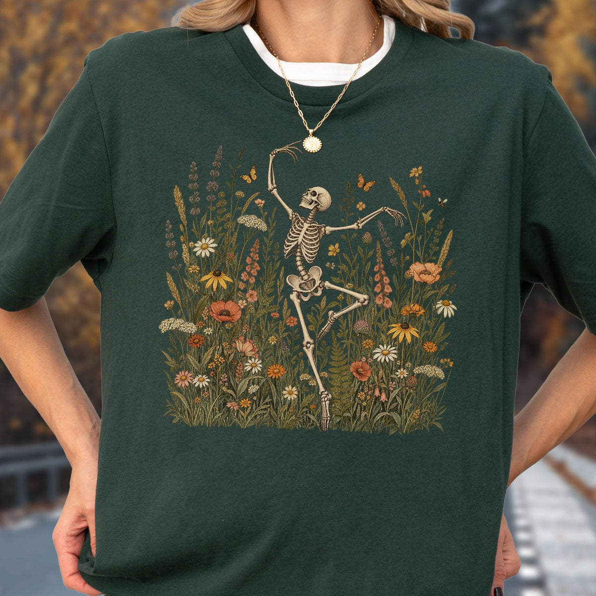 May include: A dark forest green t-shirt featuring a graphic print of a human skeleton dancing amidst a field of colorful wildflowers, including orange poppies, white daisies, and yellow coneflowers, with small orange butterflies fluttering nearby.