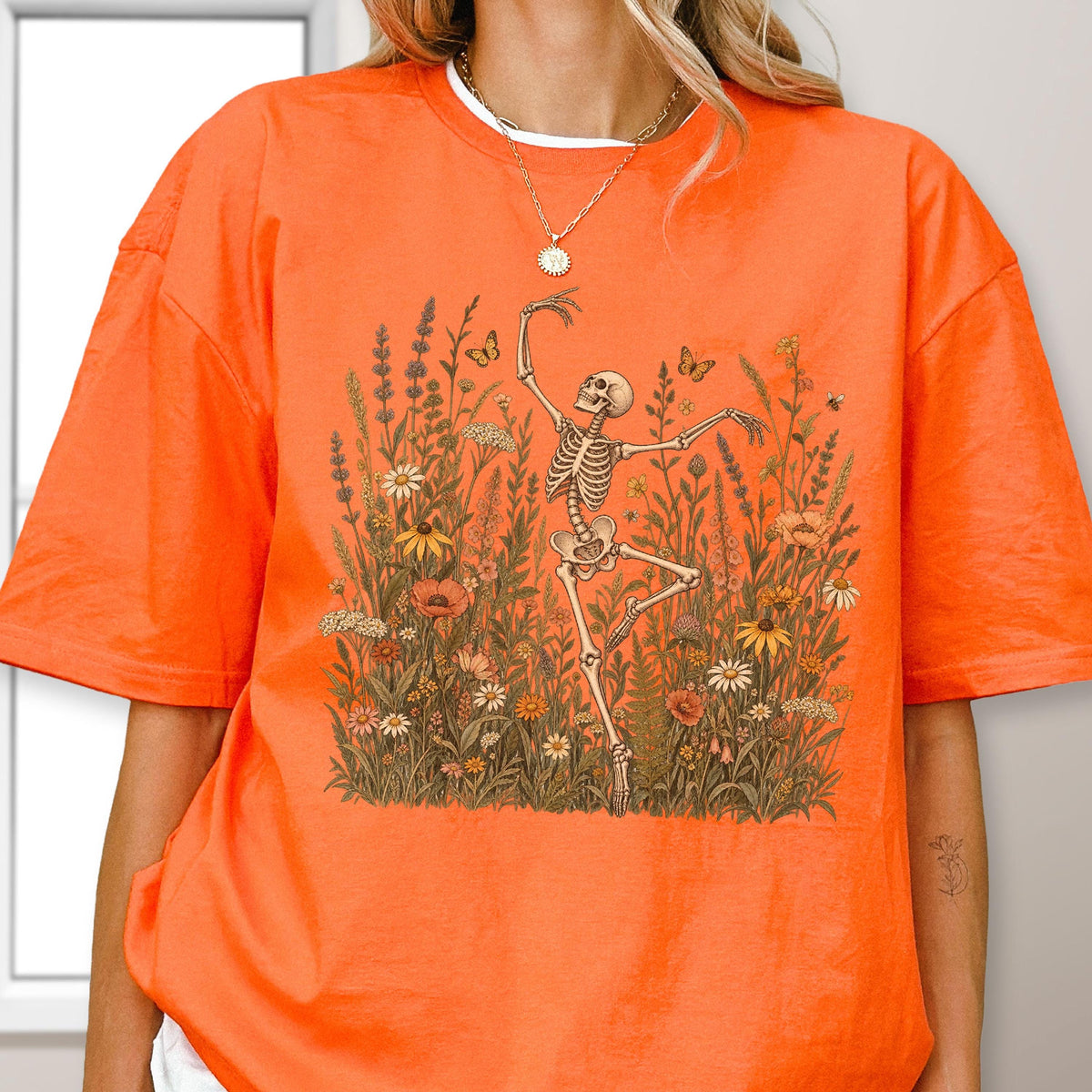 May include: A bright orange short-sleeve t-shirt featuring a graphic print of a skeleton dancing amidst a field of wildflowers, tall grasses, and small butterflies. The skeleton is positioned in the center, surrounded by various botanical elements.