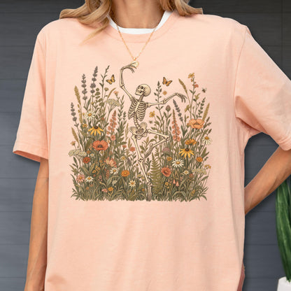 May include: A peach-colored t-shirt featuring a graphic print of a skeleton dancing amidst a field of colorful wildflowers and butterflies. The illustration includes various botanical elements like tall grasses, daisies, and poppies in muted earth tones.