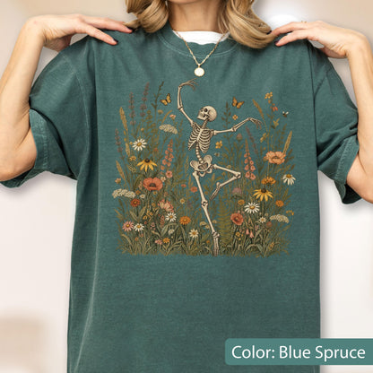 May include: A blue spruce t-shirt featuring a graphic print of a skeleton dancing amidst a field of colorful wildflowers and butterflies. The design includes various floral elements in shades of orange, yellow, and white, set against the dark fabric.