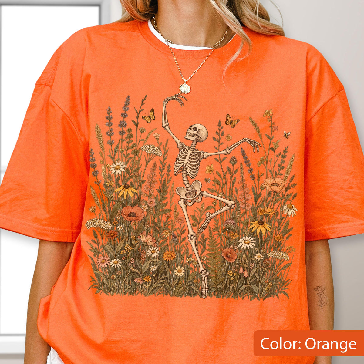 May include: An orange short-sleeved t-shirt featuring a graphic print of a skeleton dancing amidst a field of wildflowers, ferns, and butterflies. The skeleton is positioned in the center, surrounded by various botanical elements in earthy tones.