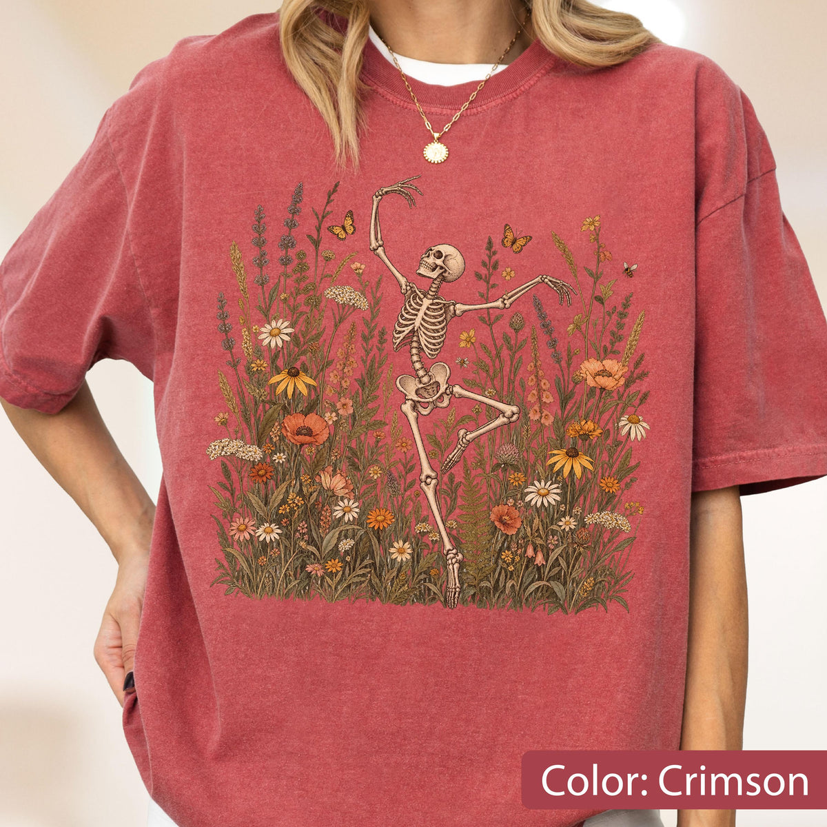 May include: A crimson short-sleeve t-shirt featuring a graphic print of a skeleton dancing amidst a field of wildflowers and butterflies. The skeleton is positioned in the center, surrounded by various orange, yellow, and white floral elements.