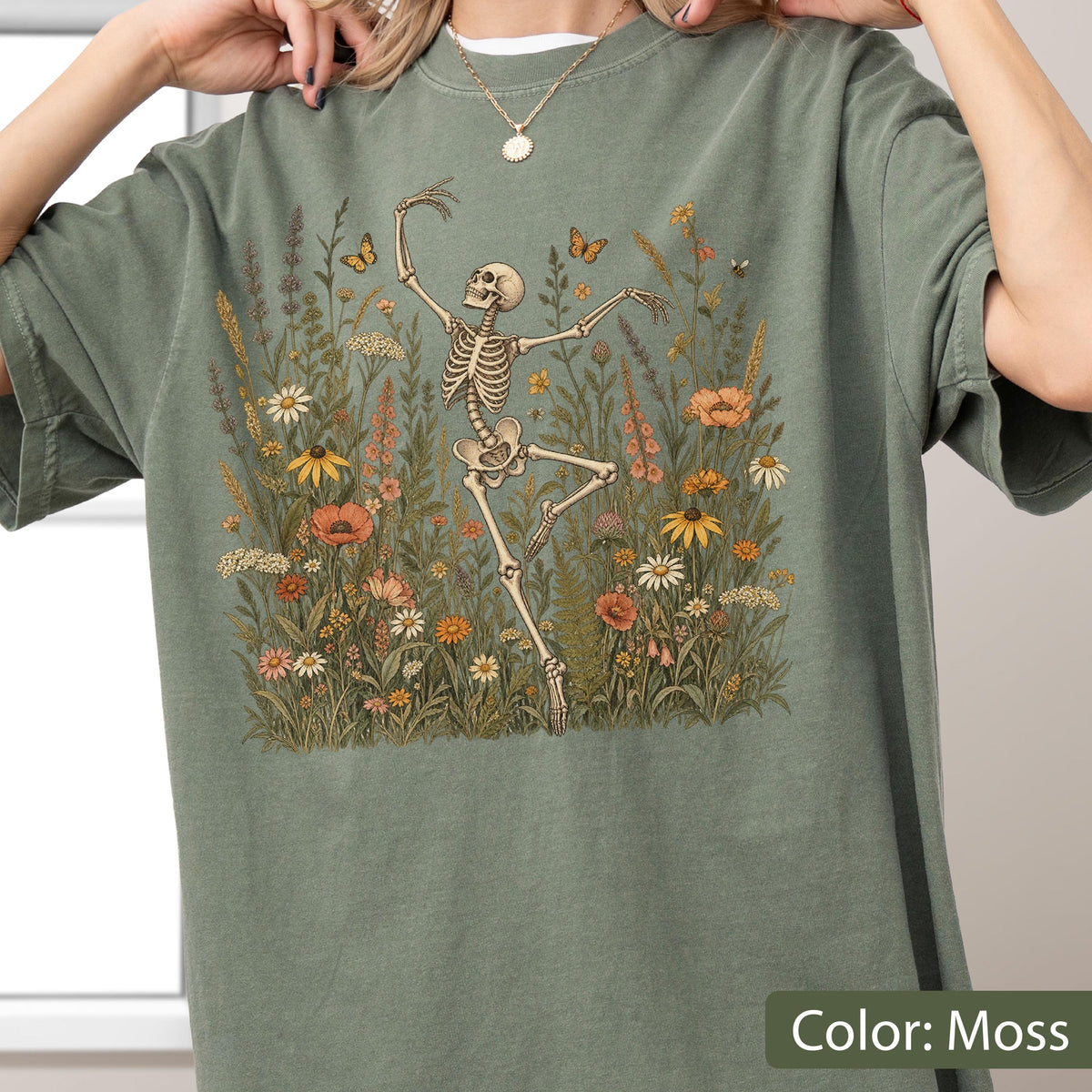 May include: A moss green t-shirt featuring a graphic print of a skeleton dancing amidst a field of colorful wildflowers and butterflies. The design includes various floral elements in shades of orange, yellow, and white, set against the muted green fabric.