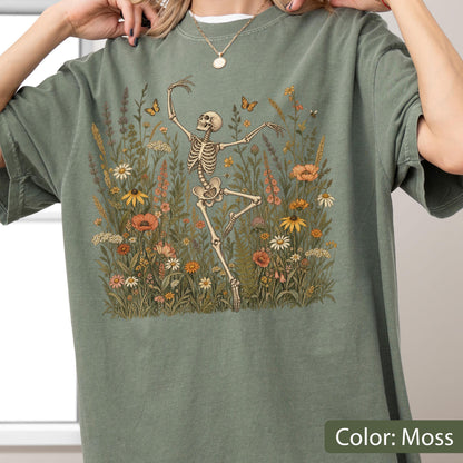 May include: A moss green t-shirt featuring a graphic print of a skeleton dancing amidst a field of colorful wildflowers and butterflies. The design includes various floral elements in shades of orange, yellow, and white, set against the muted green fabric.