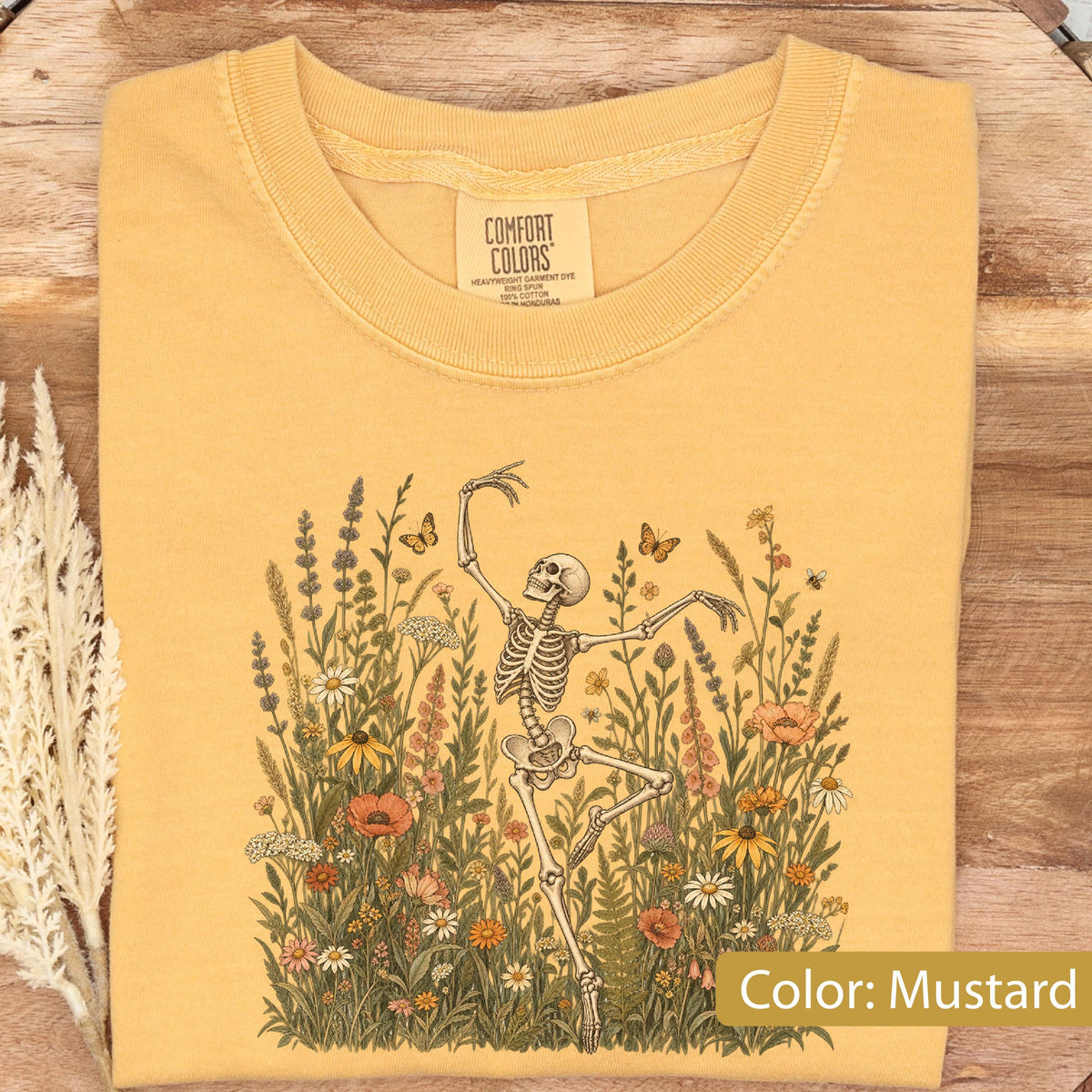 May include: A mustard yellow crew neck t-shirt featuring a graphic print of a skeleton dancing amidst a field of colorful wildflowers and butterflies. The inner neck label displays the text Comfort Colors, Heavyweight Garment Dye, Ring Spun.