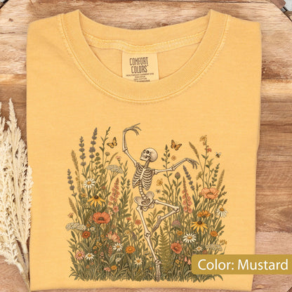 May include: A mustard yellow crew neck t-shirt featuring a graphic print of a skeleton dancing amidst a field of colorful wildflowers and butterflies. The inner neck label displays the text Comfort Colors, Heavyweight Garment Dye, Ring Spun.