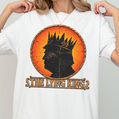 May include: A white short-sleeved t-shirt featuring a circular graphic with a distressed orange and yellow background. Inside the circle is a black silhouette of a person wearing a crown. Below the circle, the text reads THE LYING KING.