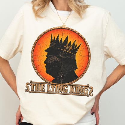 May include: A cream-colored t-shirt featuring a circular graphic with a distressed orange background. Inside the circle is a black silhouette of a person wearing a crown. Below the circle, the text reads THE LYING KING in a decorative font.