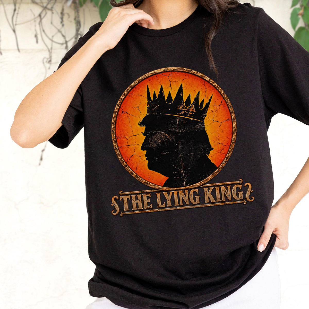 May include: A black short-sleeved t-shirt featuring a circular graphic with a distressed orange and yellow background. The graphic shows a black silhouette of a person wearing a crown, with the text THE LYING KING printed in a brown banner below.
