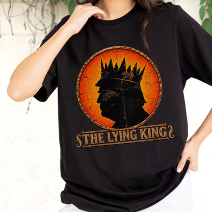 May include: A black short-sleeved t-shirt featuring a circular graphic with a distressed orange and yellow background. The graphic shows a black silhouette of a person wearing a crown, with the text THE LYING KING printed in a brown banner below.