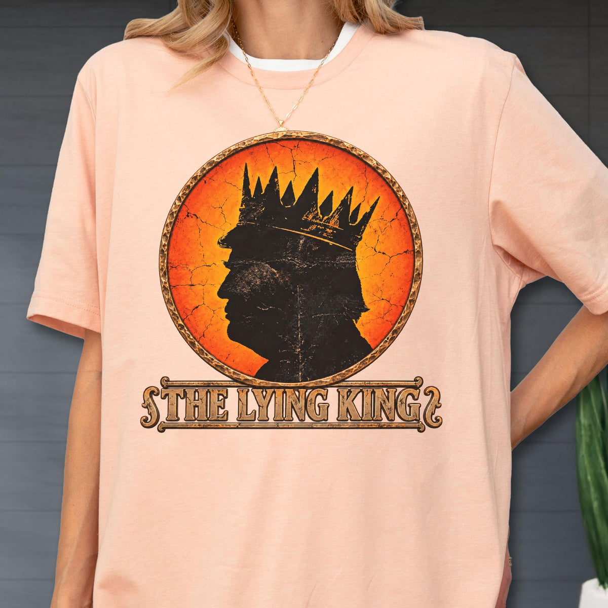 May include: A peach-colored t-shirt featuring a circular graphic with a black silhouette of a person wearing a crown against an orange and red cracked background. Below the circle, the text reads THE LYING KING in a decorative brown font.