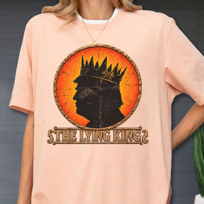 May include: A peach-colored t-shirt featuring a circular graphic with a black silhouette of a person wearing a crown against an orange and red cracked background. Below the circle, the text reads THE LYING KING in a decorative brown font.