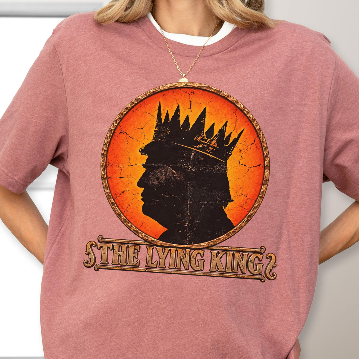 May include: A dusty rose short-sleeved t-shirt featuring a circular graphic with a black silhouette of a person wearing a crown against an orange cracked background. Below the circle, the text reads THE LYING KING in a decorative brown font.