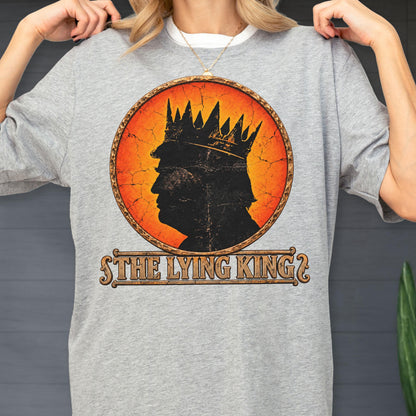 May include: A heather gray t-shirt featuring a circular graphic with a distressed orange and yellow background. The design shows a black silhouette of a person wearing a crown, with the text THE LYING KING printed in a decorative font below.