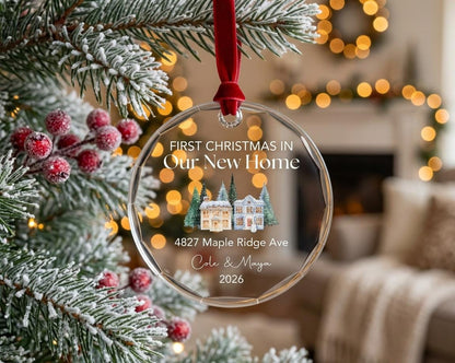 May include: A round, clear glass Christmas ornament with a faceted edge, hanging from a red velvet ribbon. It features the text "FIRST CHRISTMAS IN Our New Home", an illustration of two houses with pine trees, an address, names, and the year 2026.