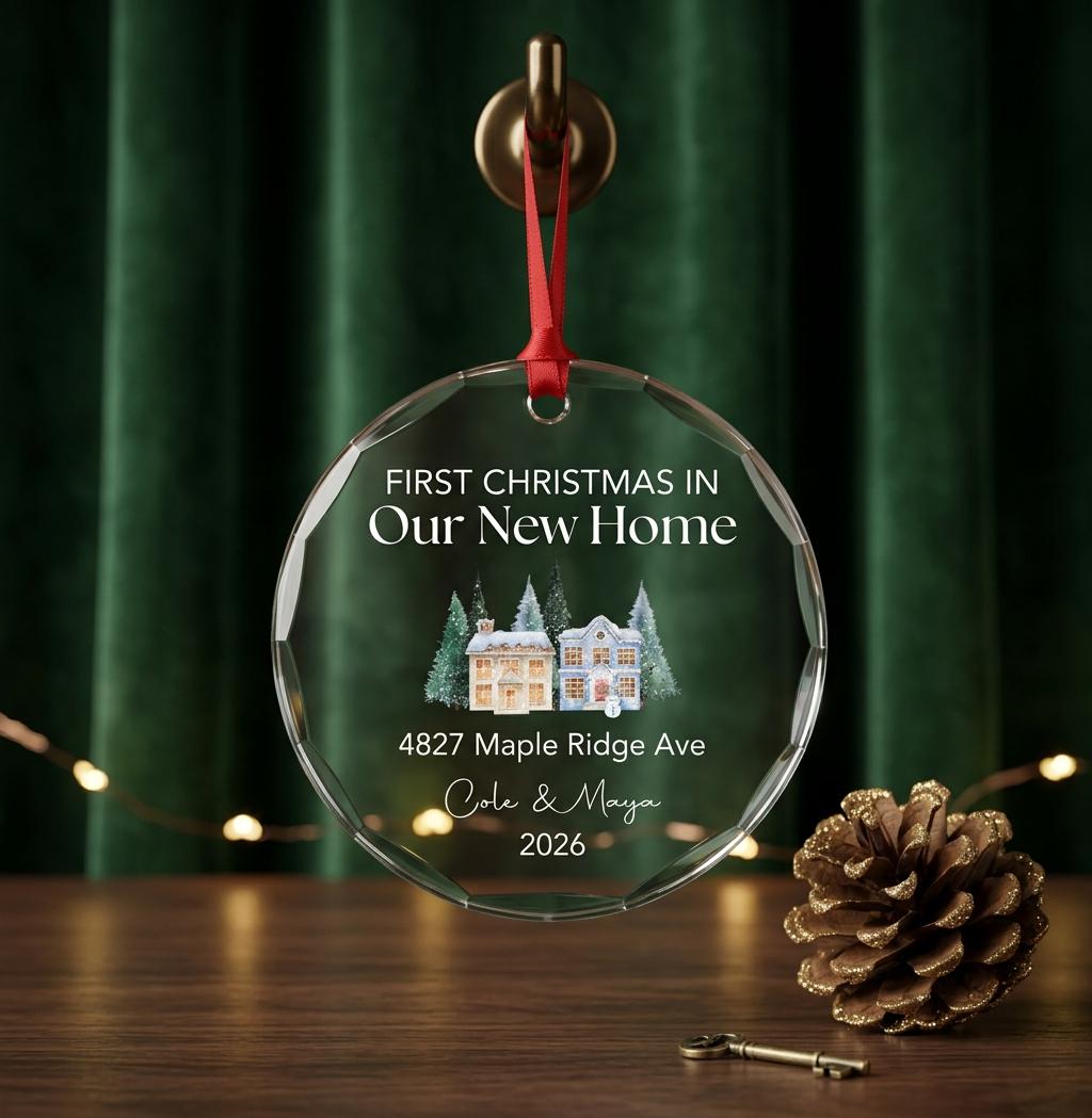 May include: A circular clear glass ornament with a red ribbon hanger. The text reads: FIRST CHRISTMAS IN Our New Home, 4827 Maple Ridge Ave, Cole & Maya, 2026. A watercolor illustration of two houses surrounded by evergreen trees is centered.