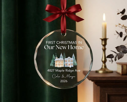 May include: A circular glass ornament with a dark red ribbon bow at the top. The text reads FIRST CHRISTMAS IN Our New Home, 4827 Maple Ridge Ave, Cole & Maya, 2026. A watercolor illustration of two houses surrounded by pine trees is centered.
