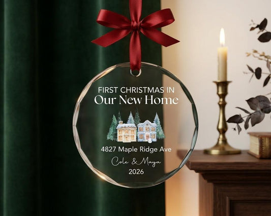 May include: A circular glass ornament with a dark red ribbon bow at the top. The text reads FIRST CHRISTMAS IN Our New Home, 4827 Maple Ridge Ave, Cole & Maya, 2026. A watercolor illustration of two houses surrounded by pine trees is centered.