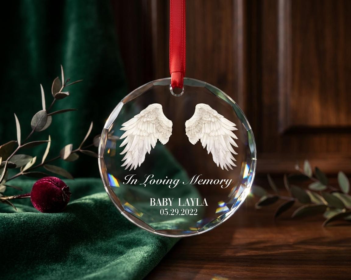 May include: A circular clear crystal ornament featuring etched white angel wings, the text In Loving Memory, the name BABY LAYLA, and the date 05.29.2022. The ornament is suspended by a thin red ribbon through a small hole at the top edge.