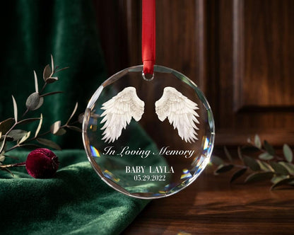 May include: A circular clear crystal ornament featuring etched white angel wings, the text In Loving Memory, the name BABY LAYLA, and the date 05.29.2022. The ornament is suspended by a thin red ribbon through a small hole at the top edge.