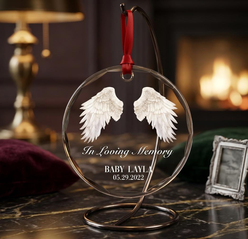 May include: A circular clear glass ornament hanging from a dark metal stand by a red ribbon. The ornament features white angel wings and the white text In Loving Memory, BABY LAYLA, and 05.29.2022 printed on the front surface of the glass.