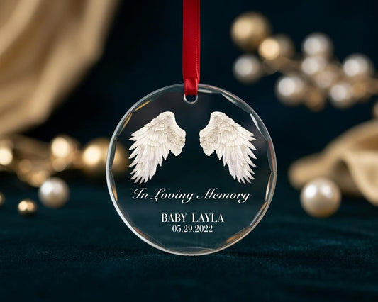 May include: A circular clear glass ornament featuring white angel wings printed in the center. Below the wings, the text reads In Loving Memory, BABY LAYLA, and 05.29.2022. The ornament is suspended by a thin red ribbon through a top hole.