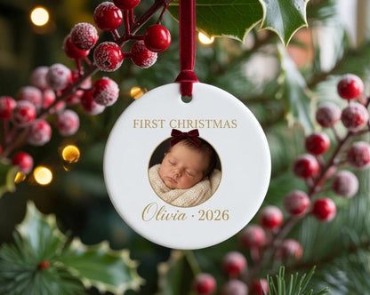 May include: A round white ceramic ornament featuring a circular photo of a sleeping infant wearing a dark red bow. The text reads FIRST CHRISTMAS above the photo and Olivia 2026 below. The ornament hangs from a dark red velvet ribbon.