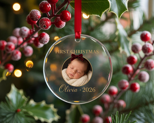 May include: A clear, circular acrylic Christmas ornament featuring a central circular photograph of a sleeping infant wearing a small dark red bow. The text FIRST CHRISTMAS is printed above the photo, with Olivia 2026 printed below in gold.