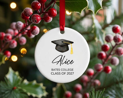 May include: A round white ceramic ornament hanging from a red ribbon. The ornament features a black graduation cap with a yellow tassel illustration, the name Alice in black script, and the text BATES COLLEGE CLASS OF 2027 in black print.