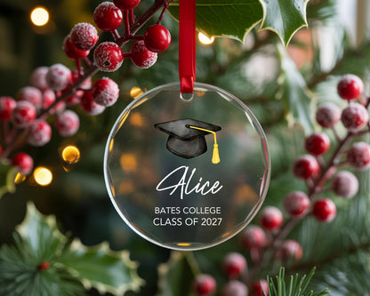 May include: A clear, circular glass ornament hanging from a red ribbon. The ornament features a black graduation cap with a yellow tassel, the name Alice in white script, and the text BATES COLLEGE CLASS OF 2027 printed in white block letters.