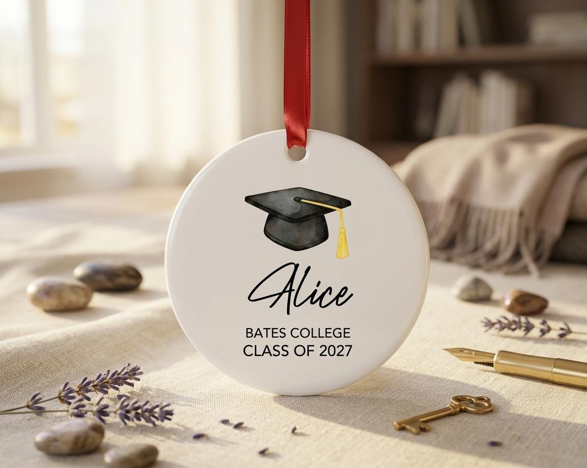 May include: A round white ceramic ornament featuring a black graduation cap with a gold tassel. The text reads Alice, Bates College, and Class of 2027. A red ribbon is attached to the top for hanging. The design is centered on the smooth surface.