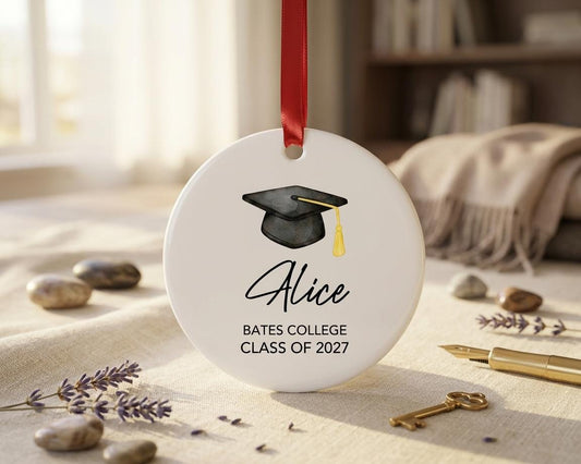 May include: A round white ceramic ornament featuring a black graduation cap with a gold tassel. The text reads Alice, Bates College, and Class of 2027. A red ribbon is attached to the top for hanging. The design is centered on the smooth surface.