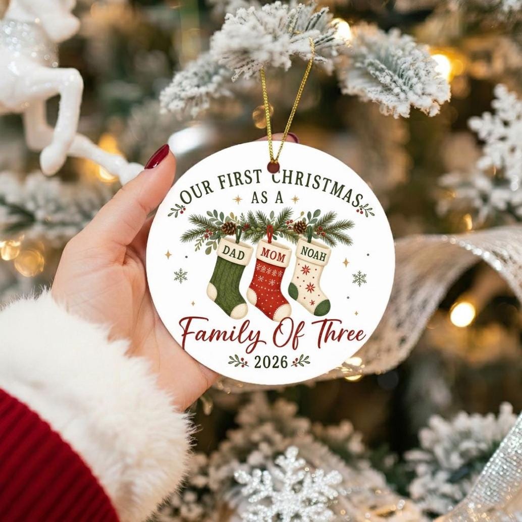 May include: A round white ceramic Christmas ornament featuring the text Our First Christmas As A Family Of Three and 2026. The design includes three hanging stockings labeled Dad, Mom, and Noah, decorated with pine branches and snowflakes.