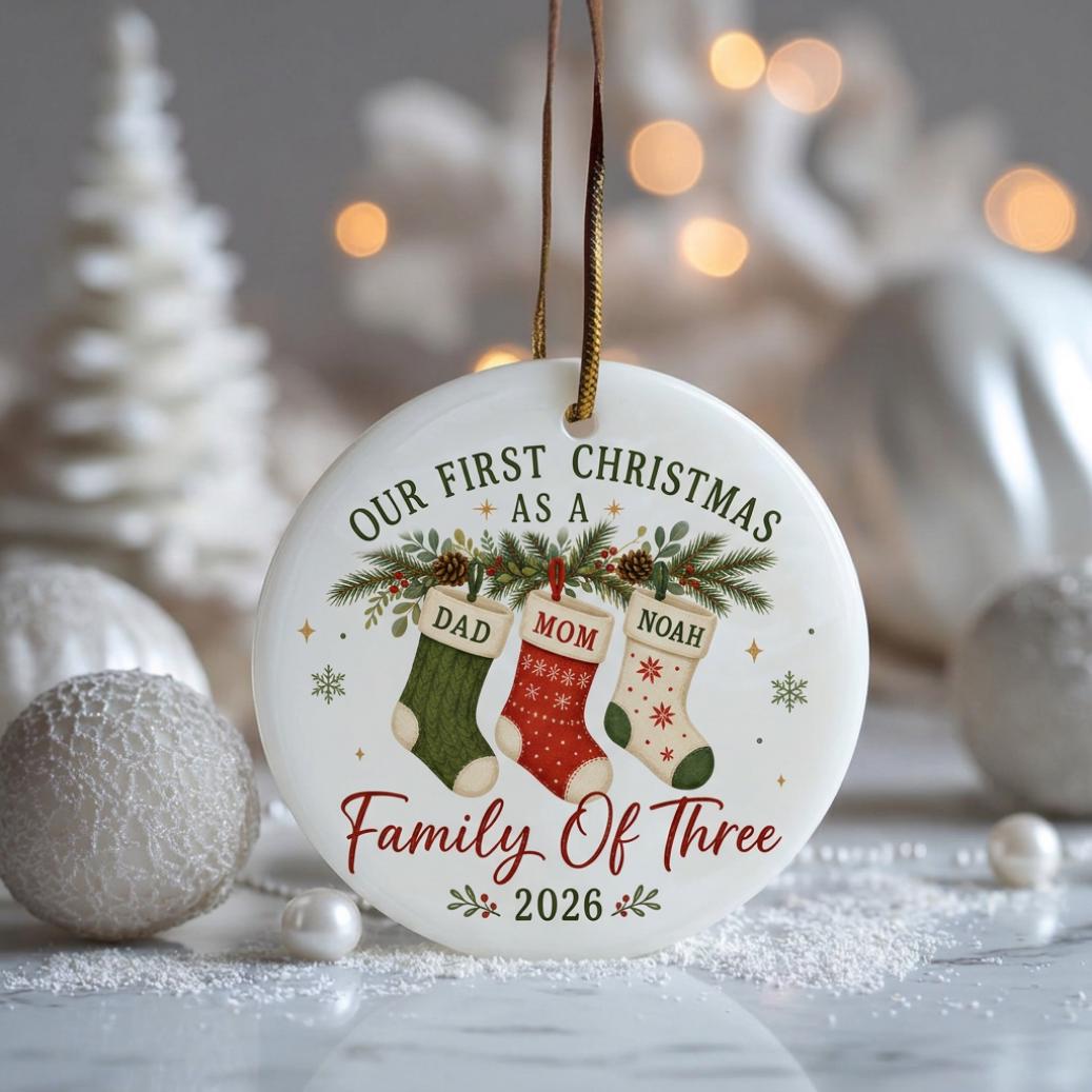 May include: A round white ceramic Christmas ornament hanging by a gold ribbon. It features the text "OUR FIRST CHRISTMAS AS A FAMILY OF THREE 2026" and an illustration of three stockings labeled "DAD", "MOM", and "NOAH" hanging from pine branches.