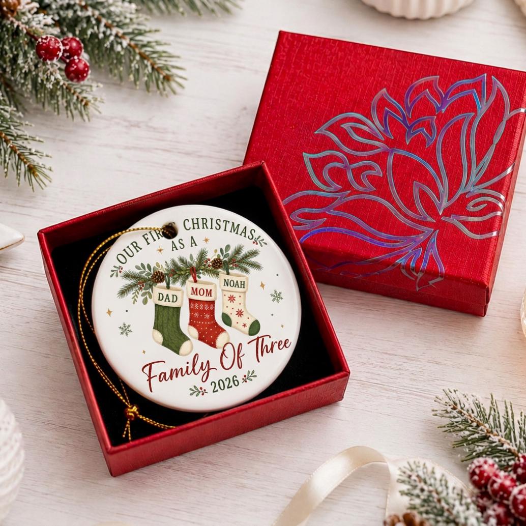 May include: A round white ceramic ornament featuring a festive illustration of three hanging stockings labeled Dad, Mom, and Noah, with the text Our First Christmas As A Family Of Three 2026, presented in a red gift box with a floral lid design.