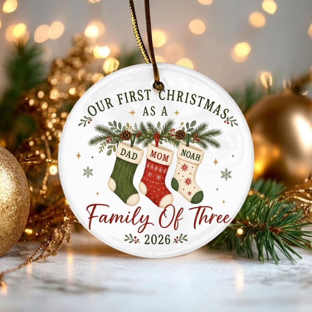 May include: A round white ceramic ornament featuring the text "OUR FIRST CHRISTMAS AS A FAMILY OF THREE" and "2026". The design includes three hanging Christmas stockings labeled "DAD", "MOM", and "NOAH" decorated with pine branches and pinecones.