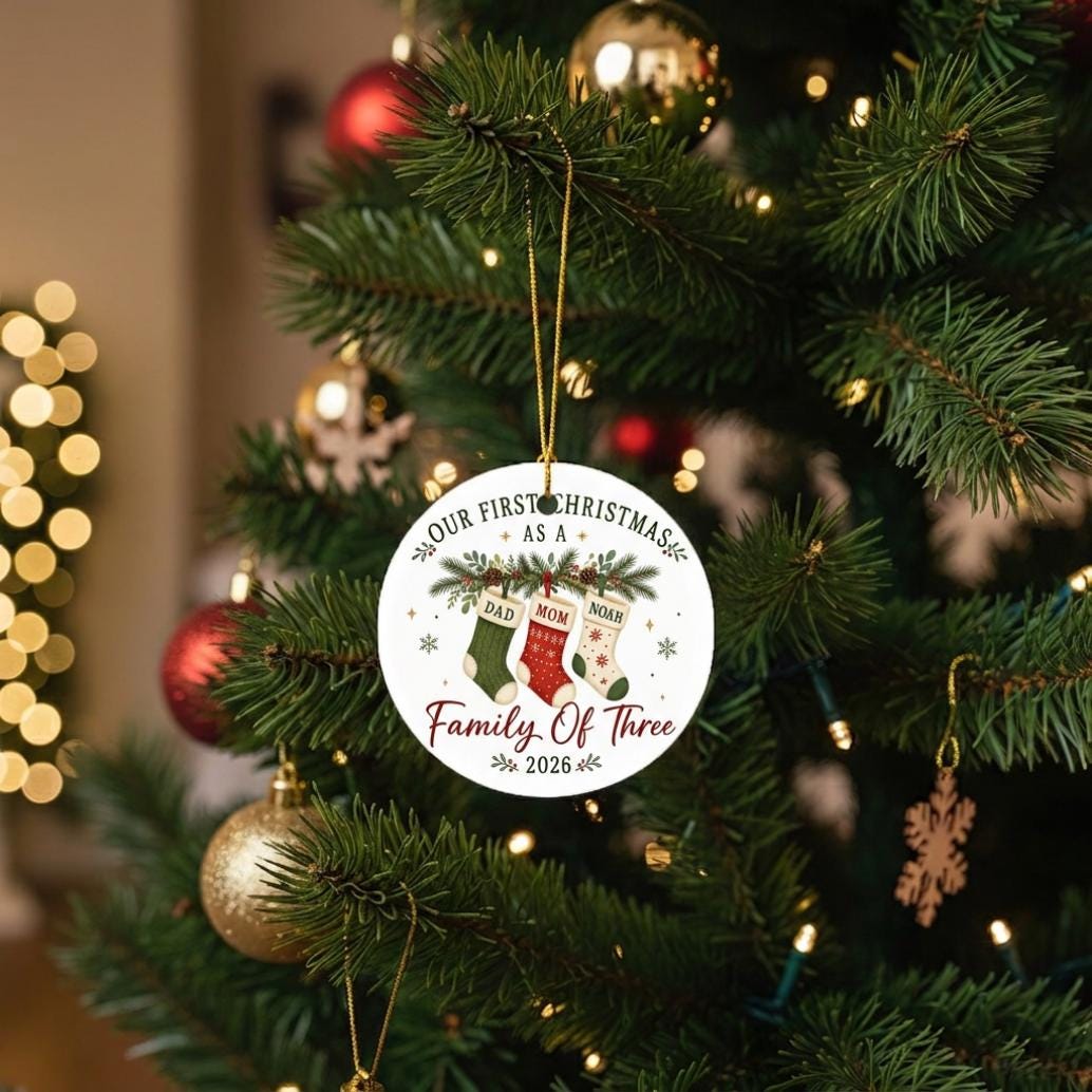May include: A round white ceramic Christmas ornament hanging by a gold string. The ornament features a graphic of three stockings labeled Dad, Mom, and Noah, with the text Our First Christmas As A Family Of Three 2026 printed in black and red.