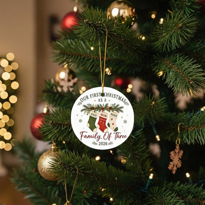 May include: A round white ceramic Christmas ornament hanging by a gold string. The ornament features a graphic of three stockings labeled Dad, Mom, and Noah, with the text Our First Christmas As A Family Of Three 2026 printed in black and red.