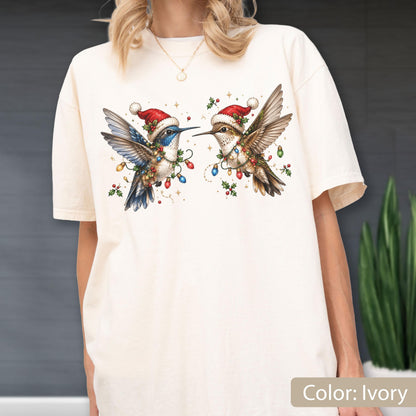 May include: An ivory t-shirt featuring a graphic of two hummingbirds wearing red Santa hats. The birds are decorated with colorful Christmas lights and holly sprigs. The design is centered on the chest of the shirt, showcasing detailed feathers and festive holiday accents in red, green, blue, and yellow hues.