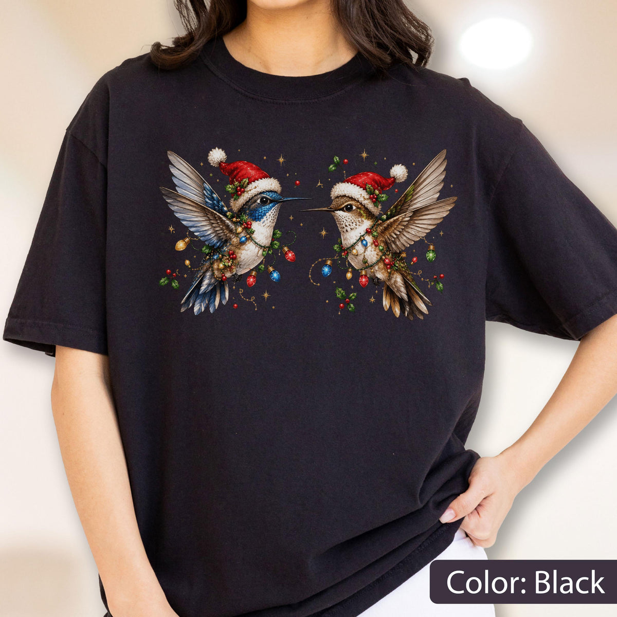 May include: A black t-shirt featuring a graphic of two hummingbirds facing each other. Both birds wear red and white Santa hats and are surrounded by colorful Christmas lights, holly berries, and green leaves. The design is centered on the chest.