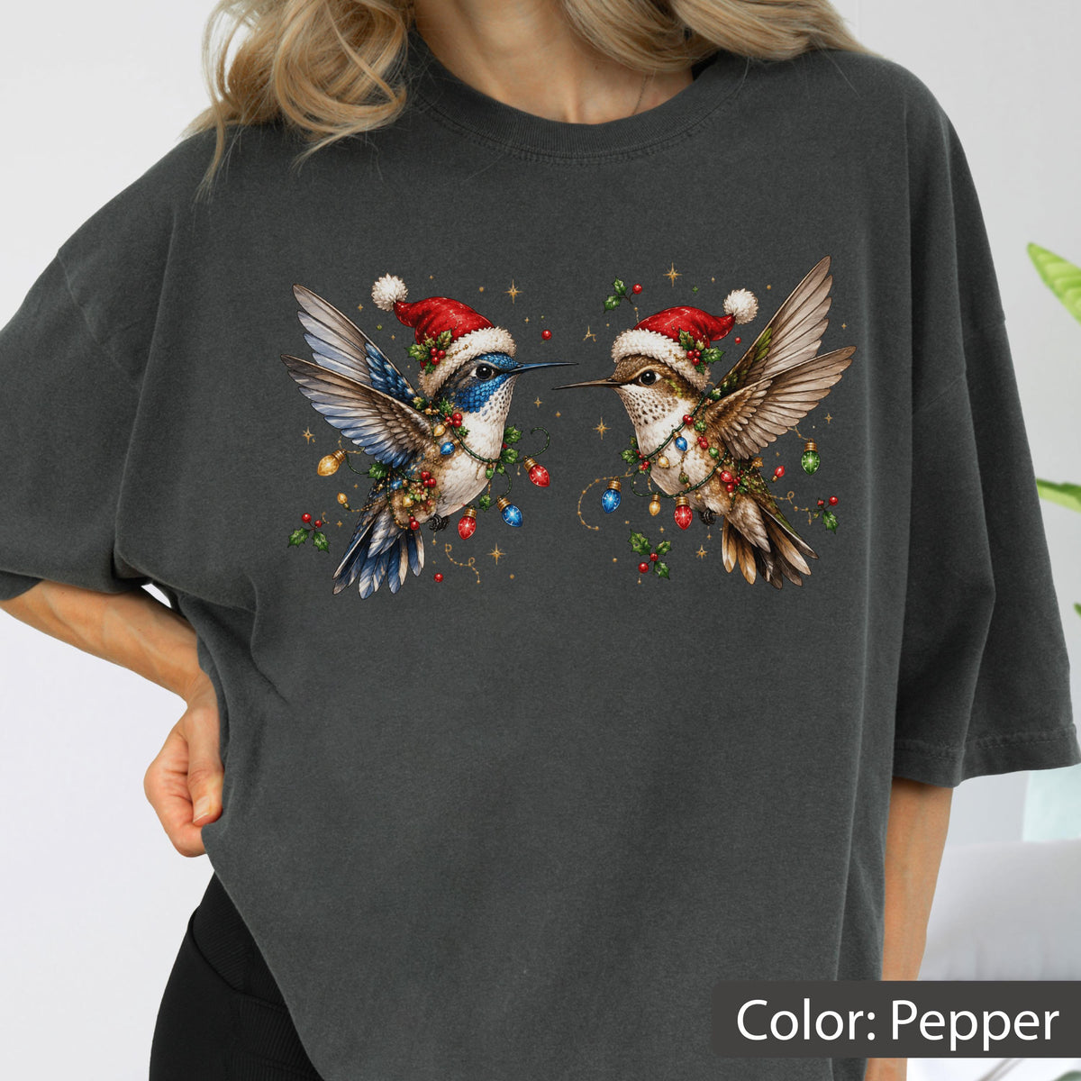 May include: A charcoal gray t-shirt featuring a graphic of two hummingbirds wearing red and white Santa hats. The birds are decorated with colorful Christmas light bulbs and holly berries, set against a background of small golden stars.