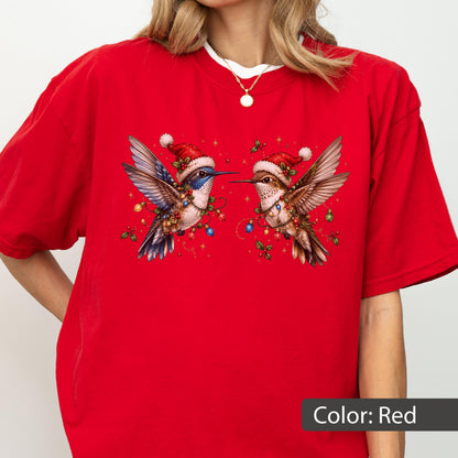 May include: A bright red short-sleeved t-shirt featuring a graphic of two hummingbirds facing each other. Each bird wears a red Santa hat and is wrapped in colorful Christmas lights with green holly leaves and small gold stars in the background.
