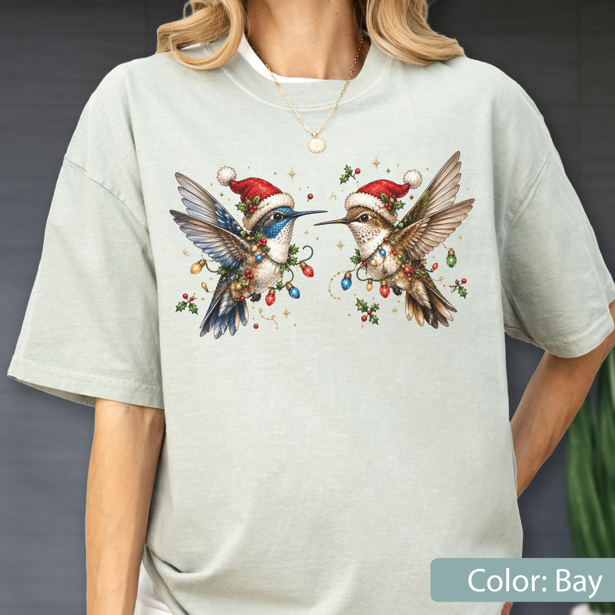 May include: A light sage green t-shirt featuring a graphic of two hummingbirds wearing red Santa hats. The birds are surrounded by colorful Christmas light bulbs and holly berries. The design is centered on the chest of the short-sleeved garment.