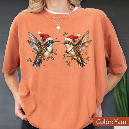 May include: A short-sleeved crew neck t-shirt in a solid yam orange color. The front features a graphic illustration of two hummingbirds facing each other, both wearing red Santa hats and wrapped in colorful Christmas light strings and holly.